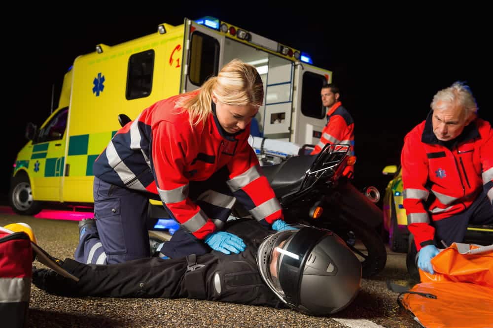 Image is of paramedics providing roadside care to an injured rider, showing emergency response in a motorcycle crash case.