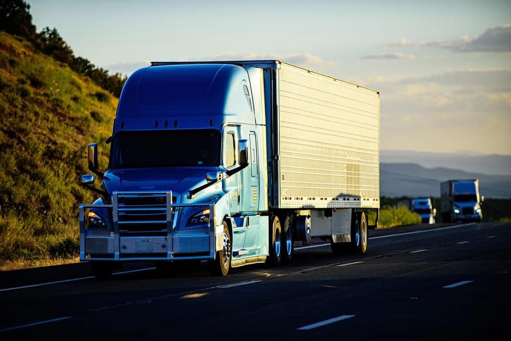Image is of a semi truck driving on a highway during daylight, showing conditions that can contribute to truck accidents caused by long hours on the road