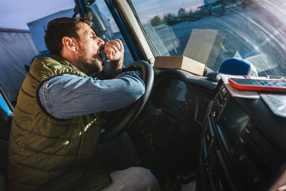 Image is of a truck driver yawning while driving inside the cabin, demonstrating how fatigue and drowsiness increase the risk of truck accidents