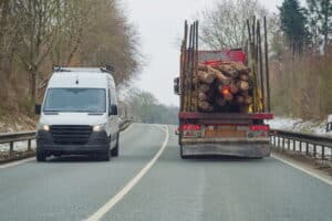 Image is of a logging truck carrying timber beside another vehicle, concept of cargo spill risks on Tennessee roads.
